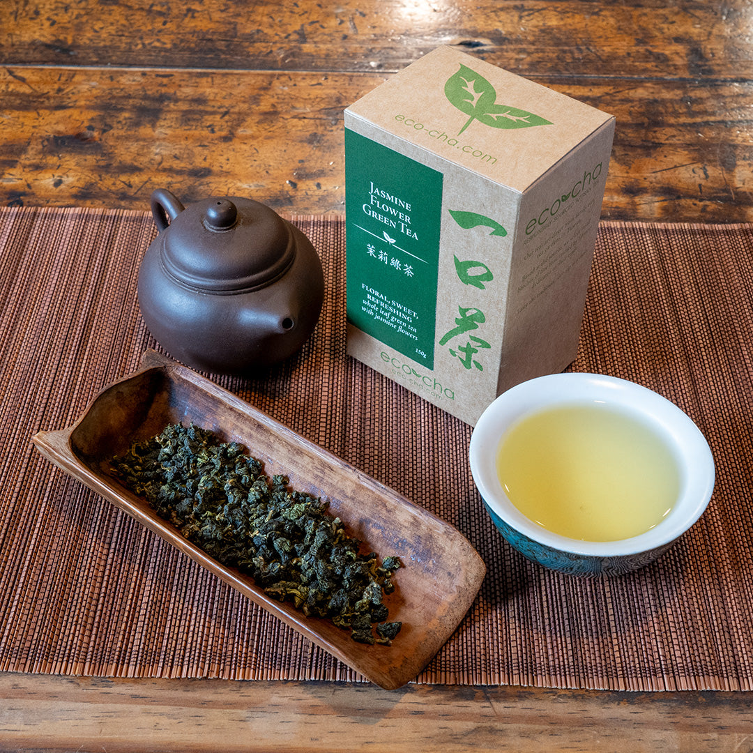 Jasmine Flower Green Tea in a tea scoop next to a brown tea pot and box packaging. A blue and white tea cup is filled with brewed tea.