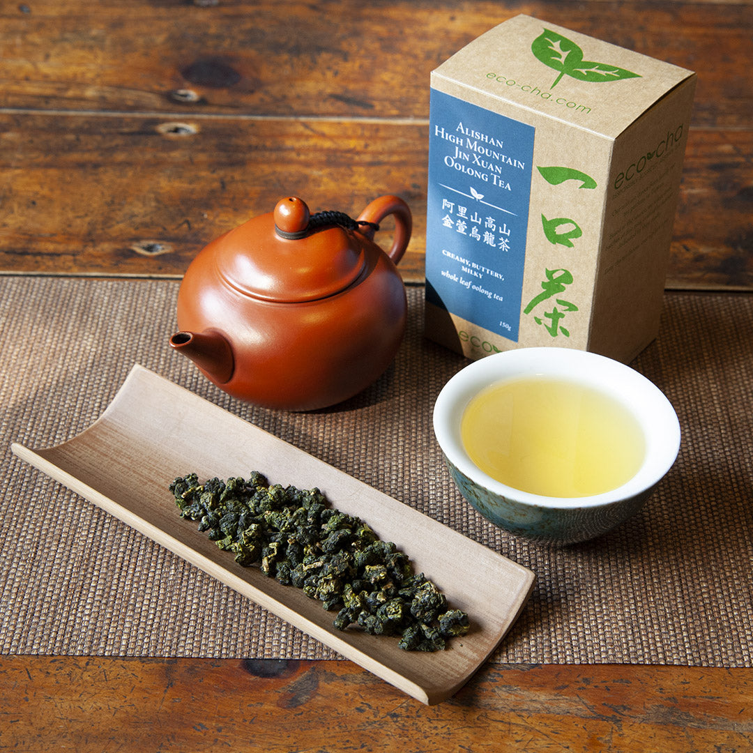 Alishah High Mountain Jin Xuan Oolong Tea dry tea leaves on a bamboo scoop on a dark wooden table. Red teapot and brewed tea in a cup beside it. Packaged tea in a box in the background.