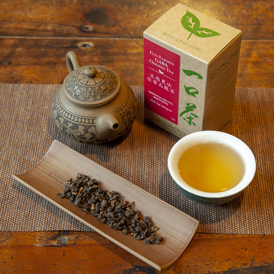 Eco-Farmed GABA Oolong Tea in a cup on a wooden table next to dry leaves and a teapot and a box
