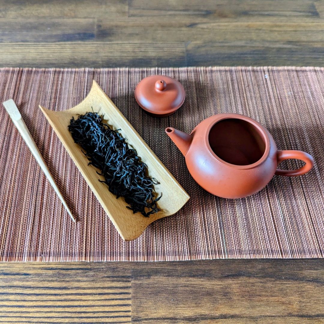 Bamboo tea scoop and tea pick next to a red clay teapot on a woven mat