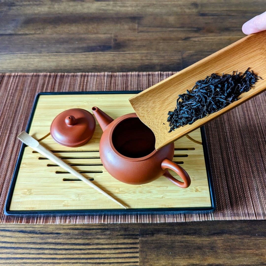 Black tea leaves being poured into a red clay teapot with a bambook tea scoop