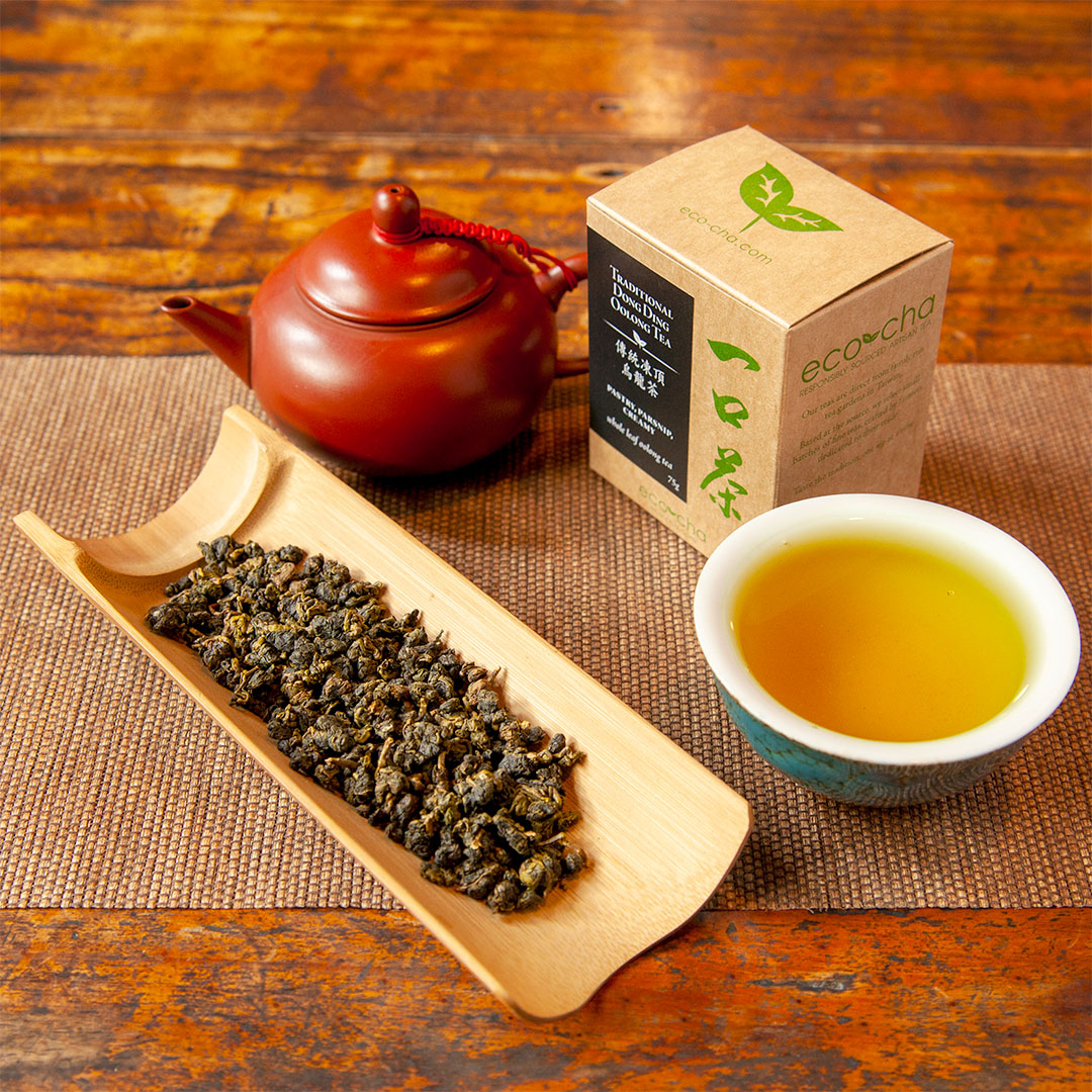 Eco-Cha Teas Traditional Dong Ding Oolong Tea dry leaves on a bamboo scoop in front of a red clay teapot and a cup of brewed tea and a packaging box.