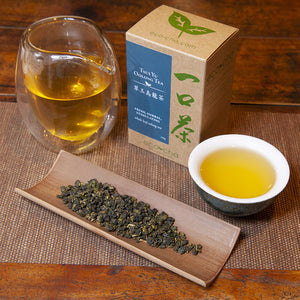 Tsui Yu Oolong Tea in a teacup on a wooden table next to dry tea leaves, a pitcher of tea, and a box