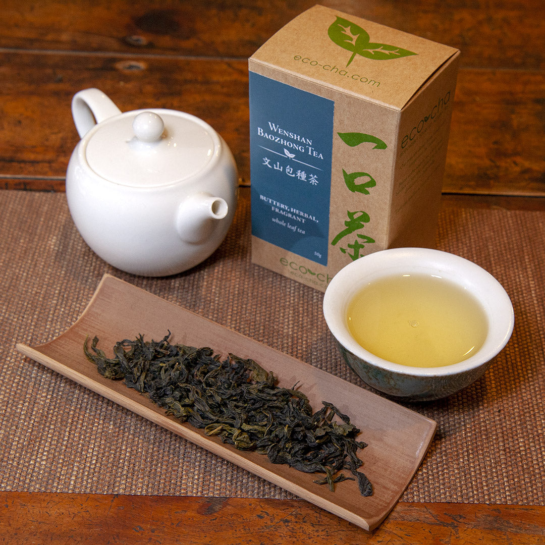 Wenshan Baozhong Tea in a teacup on a wooden table next to dry tea leaves and a white teapot and box